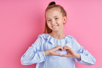happy caucasian girl shows heart made from hands at camera, positive child in blouse smiles, looks a