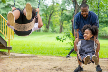 Happy affectionate mixed race family. African man father with two little daughter playing swing together at playground. Dad and child girl enjoy and having fun together in outdoor weekend vacation.