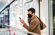 © Sanja - Young handsome fashionable man with protective face mask on standing in shopping mall, carrying bags and using phone during corona virus outbreak.