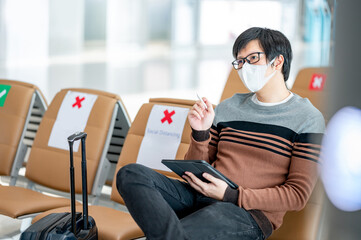 Wall Mural - Asian man tourist with suitcase luggage wearing face mask using digital tablet in airport terminal. Coronavirus (COVID-19) prevention when travel abroad. Health awareness and social distancing concept