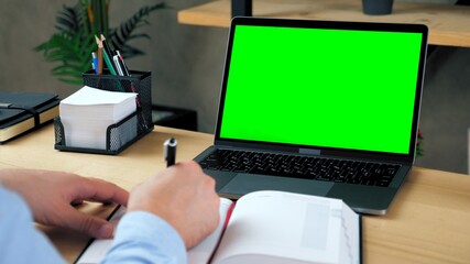 Man student at home office sits table study writes information with pen in notebook. Green screen laptop computer concept: online remote distance learning education by web cam video call conference