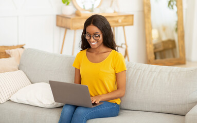 Wall Mural - Portrait of young black lady using laptop computer for work or education on couch at home