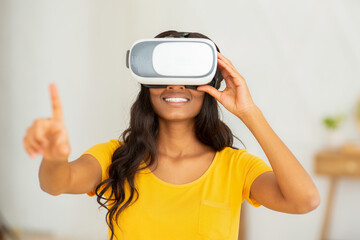 Wall Mural - Modern technologies concept. Positive young black woman in VR headset pushing imaginary button in cyberspace at home