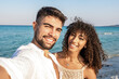 Beautiful mixed race couple taking a selfie at the sea at dusk - Handsome young man in white open shirt with his latina curly haired girlfriend smiling together in summer vacation