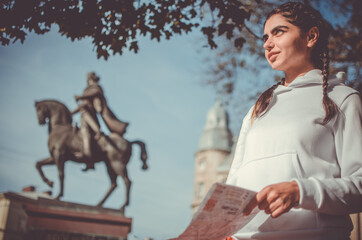 Young cute smiling girl with braids in white hoodie with map in her hands under the tree near the monument on a sightseen tour