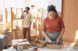 © AnnaStills - Young woman packing parcels into the boxes at the table with her colleague talking on mobile phone in the background they working in delivery service