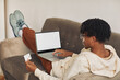 © AnnaStills - Rear view of African man lying on sofa with laptop he working online and using credit card for paying the bills