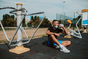 Wall Mural - A fit muscular male caucasian athlete with a face mask sits next to a equipment in an outdoor gym and drinks water from a bottle. Coronavirus protection COVID - 19