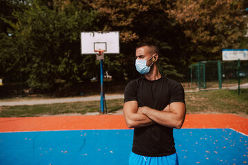 Wall Mural - Profile of a young muscular male caucasian athlete with a face mask standing on an outdoor basketball court. Coronavirus protection COVID - 19