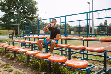 Wall Mural - A fit young male caucasian athlete with a face mask sitting on the stands of a football field and talking on the phone. COVID - 19 coronavirus protection