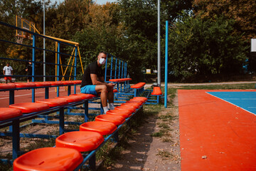 Wall Mural - A young muscular male caucasian athlete with a face mask sitting on the tribune of a football field and texting message on the phone. COVID - 19 coronavirus protection