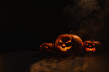 Three Creepy Halloween Steaming Pumpkins With A Carved Luminous Smirk On A Black Background. A Handmade Jack-o-lantern Head With A Candle Inside In The Dark Among The Fog. Trick Or Treat. Postcard.