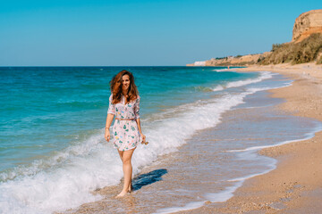 Beautiful smiling woman in a dress walking on the sand along the emerald sea, leisure and tourism concept