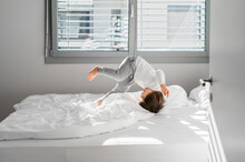 Happy Little Child Boy Jumping On Bed Doing Somersault Having Fun At Home At Sunny Morning.
