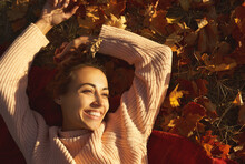 Carefree Woman Lying On Fallen Leaves And Enjoying Warm Sunny Weather At Autumn Park.