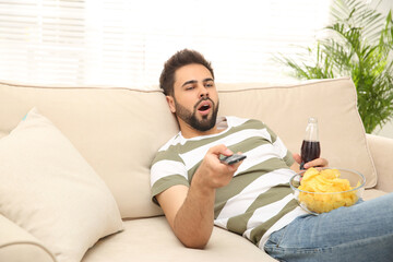 Lazy young man with chips and drink watching TV on sofa at home