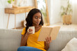 © Prostock-studio - Stay home hobbies concept. Attractive millennial black lady reading book with cup of hot beverage in living room
