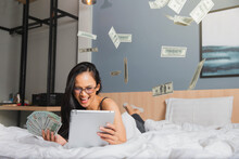 Young Woman Counting A Lot Of Dollar Notes She Has Made A Profit From The Investment. Happy Woman Counting Dollars Cash On Bed. Woman Laying On Bed Enjoying Use Counting Dollars Cash With A Laptop.
