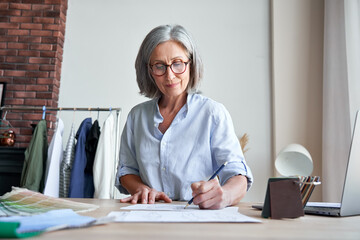 Smiling middle aged stylish woman fashion designer drawing sketches in studio. Mature old adult elegant grey-haired lady dressmaker small business owner creating new fashion design cloth in atelier.