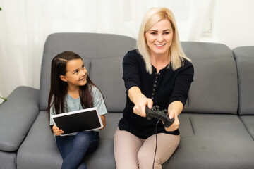 good relationship cute little girl with young mother using joystick playing video game sitting together in living room enjoying family holiday.