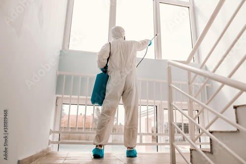 Worker in white sterile uniform, with rubber gloves and mask on holding sprayer with disinfectant and sterilizing windows in school..