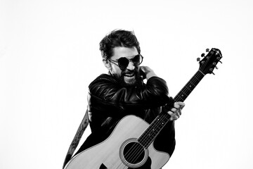 A man plays the guitar in a black leather jacket with sunglasses on a light background