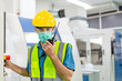 © winnievinzence - Male worker in surgical face mask due to covid19 pandemic talking on radio phone or walkie-talkie with colleague to control work in front of technology machine at industrial plant factory. copy space