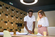 Content African American designer writing with pencil on draft and explaining details to client. Pretty blonde woman standing near table and listening guy. Communication, startup and teamwork concept