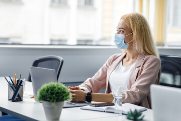 Wall Mural - Business must be continuous, healthcare concept. Blond woman in protective mask at workplace with laptop and antiseptic and smartphone looks out the window in interior