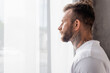 © LIGHTFIELD STUDIOS - side view of bearded, tattooed man looking away while standing near window at home