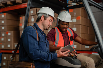 Wall Mural - Two male workers wearing hard hats holding digital tablet researching location of parcel while driving factory card in warehouse.