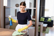 chambermaid in apron and rubber gloves holding spray bottle and rag while cleaning wooden surface in hotel room