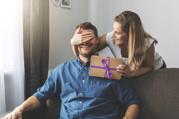woman giving surprise gift to beloved man at home. closed eyes with hand from behind