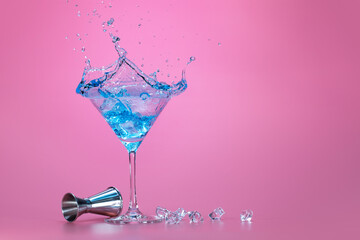 Wall Mural - blue liquid splashing in a martini cup, jigger and fragments of ice cubes isolated on pink background with space for text