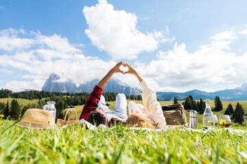 Wall Mural - Romantic couple in love doing picnic visiting mountains alps. Boyfriend and girlfriend enjoying love doing heart shape with hands outdoor.