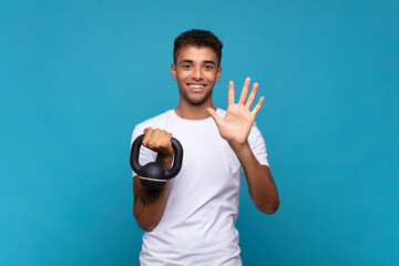 Wall Mural - young man lifting a sumbbell smiling and looking friendly, showing number five or fifth with hand forward, counting down