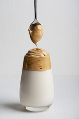 One drinking glass full of trendy dalgona coffee made by whipping instant coffee powder, sugar and hot water with addition of cold milk on white wooden background with spoon making cap. Vertical