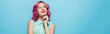 dreamy young woman with pink hair smiling and looking away isolated on blue, panoramic shot