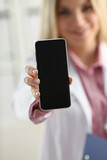 Fototapeta  - Beautiful female blonde doctor holding smartphone in hand at office woman discussing disease and giving an online consultation to a remote training mobile advertising app