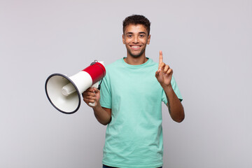 Wall Mural - young man with a megaphone smiling and looking friendly, showing number one or first with hand forward, counting down