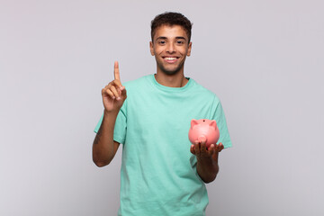 Wall Mural - young man with a piggy bank smiling and looking friendly, showing number one or first with hand forward, counting down