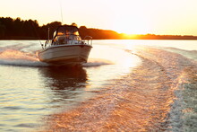 Fast Gliding Motor Boat In Wake Track On Beautiful Sunset Background, Bright Orange Sun Shine In Calm Water On Forest Stripe On Horizon At Sunny Summer Evening, Front Side View From Powerboat Stern