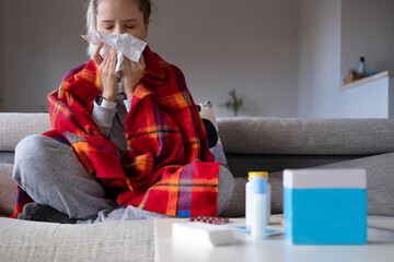 Sick runny nose woman wrapped up on scarf with handkerchief trying to health with medicine and pills on home backdrop. Health care concept. Coronavirus, disease prevention. Self isolation.