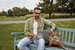 © Kostiantyn - Young handsome man sitting on the wooden bench in park and using laptop, working outdoors