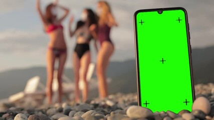 Phone with a green screen and tracking dots on the beach, three multi-racial girls in swimsuits dancing in the background. Chromakey