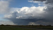 Time Lapse Of A Large Billowing Thunder Cloud With Rain Falling.  A Gravel Road Cuts Through The Foreground And Autumn Grass And Trees Make Up The Mid-ground. 
