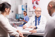 © DC Studio - Senior medical practitioner taking notes on clipboard during consultation of married couple in hospital office. Doctor looking at male patient while wearing white coat and stethoscope.