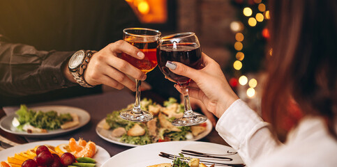 hands with glasses clinking against the background of Christmas tree lights and bonfires from a home fireplace over a table with delicious dishes