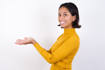 Young hispanic girl with short hair wearing casual yellow sweater isolated over white background pointing aside with hands open palms showing copy space, presenting advertisement smiling excited happy