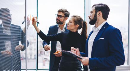 Side view of Caucasian colleagues drawing diagram on modern glass board for planning business strategy, collaboration of experienced male and female partners during working process in office interior